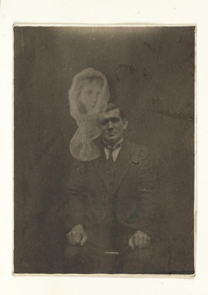 Black and white phpotograph of a seated man showing a spectral female head above him