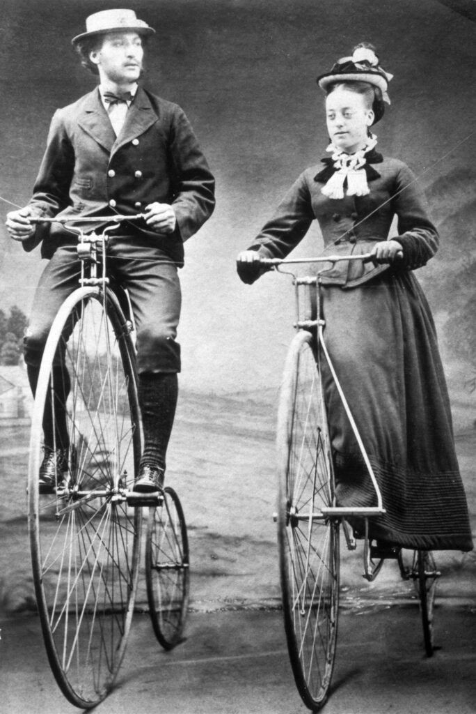 Monochrome photograph of a man and a woman riding penny farthing bicycles