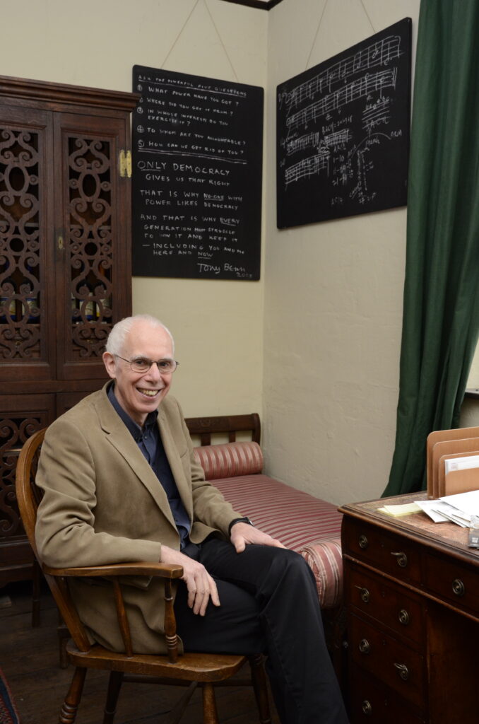 Colour photograph of Jim Bennett sat at desk with blackboards hung behind