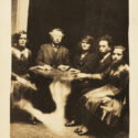 Image of five people sitting around a Oujia board. One of them has glowing eyes and a hand seems to extend toward him from the floor. This is produced by photographic manipulation. The photograph is set against an ivory background.