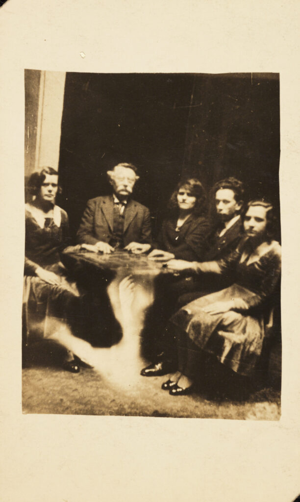 Image of five people sitting around a Oujia board. One of them has glowing eyes and a hand seems to extend toward him from the floor. This is produced by photographic manipulation. The photograph is set against an ivory background.