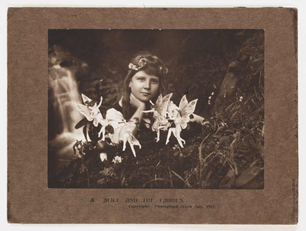 Image of a scanned photograph in sepia tones of a young girl playing with fairies. Caption printed on the brown carboard background says 'A. Alice and the Fairies'