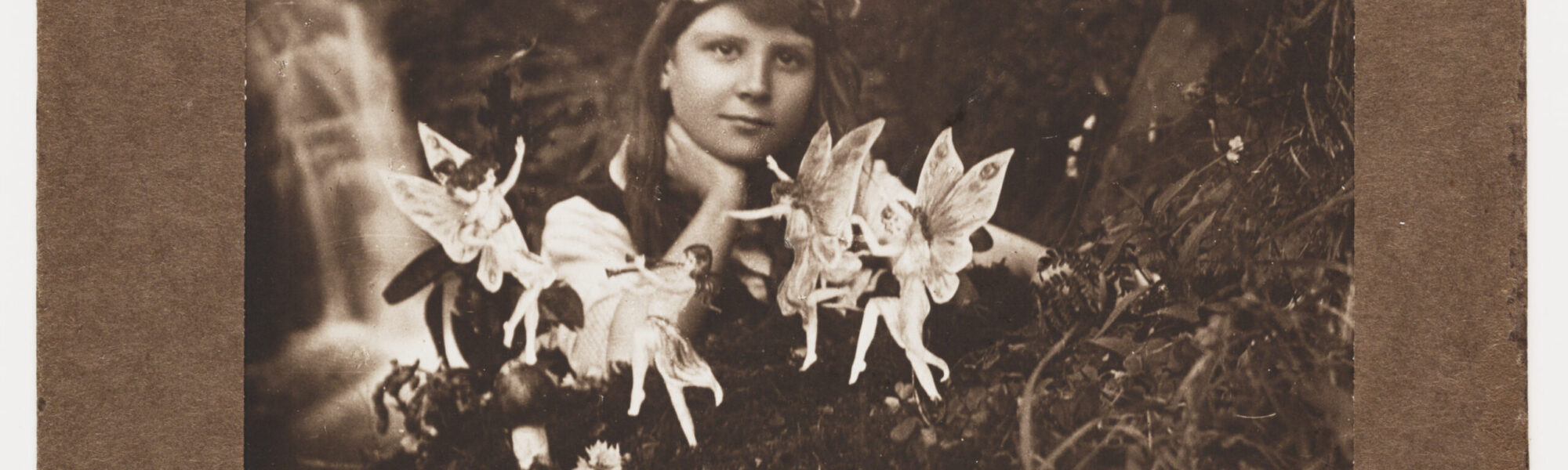 Image of a scanned photograph in sepia tones of a young girl playing with fairies. Caption printed on the brown carboard background says 'A. Alice and the Fairies'