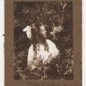 Photograph of a young girl turned to her left looking at a fairy. She is wearing a white dress and white bow. The fairy is two-dimensional and has been made out of paper. They are set against a woodland background. The photograph has been pasted to a brown cardboard background, on which has been printed 'C. Alice and Leaping Fairy'