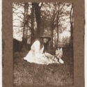 Scanned photograph of a young girl outside in the grass with trees in the background. The photograph shows her in a white dress with a hat playing with a whimsical woodland creature, identified as a gnome by the caption. The caption 'B. Iris and the Gnome' has been printed on the cardboard.