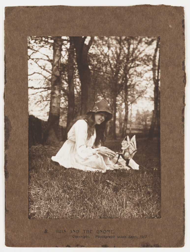 Scanned photograph of a young girl outside in the grass with trees in the background. The photograph shows her in a white dress with a hat playing with a whimsical woodland creature, identified as a gnome by the caption. The caption 'B. Iris and the Gnome' has been printed on the cardboard.