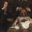 Image of an anatomy lesson taking place. In the center is a corpse with skin open at the skull so that the brain is on full display. To the left is a man watching attentively, dressed in black coat and white shirt. Behind is the torso of a man holding tools as he cuts into the brain.