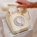 Image of a white telephone, with its receiver being picked up by the hand of someone off-camera.