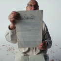 A man in a white shirt holds up a paper that reads 'This Indenture', wood shards in the background.