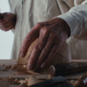 A man's hand holding wood, with carving tools in the foreground.