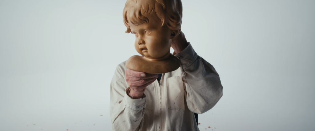 A man holds up a cherubic head sculpted out of wood. Behind, the viewer can see his torso in a white shirt.