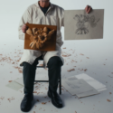 A man holds up a sketch and a wood carving next to each other. He wears a white shirt and black trousers and sits in a chair. In the background are wood shavings and other discarded sketches.