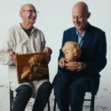 Two men sit next to each other, one holding a carved cherubic wooden head and the other a sculpture of a floral wooden rectangle