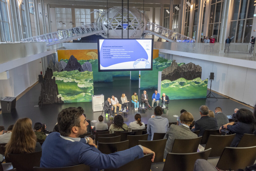 A number of people sitting in front of a discussion group that stands on stage. There is a large screen behind the discussion group, as well as various environment-themed additional decorations.