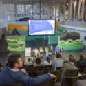 A number of people sitting in front of a discussion group that stands on stage. There is a large screen behind the discussion group, as well as various environment-themed additional decorations.