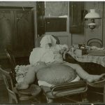 Black and white photograph of an adult male showing elephantiasis of the scrotum wearing a towel as a hood to cover the face