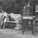 Black and white photograph of Beatrice Harrison playing the cello in her garden seated next to a dog and surrounded by pet birds in cages