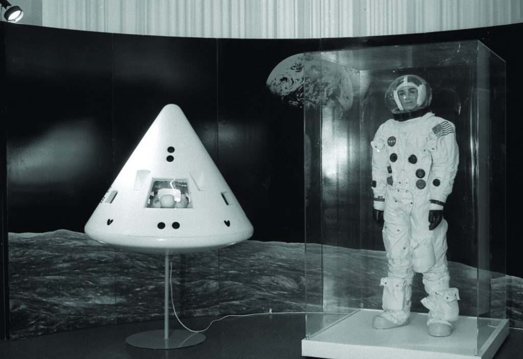 Black and white photograph of an exhibition display containing a spacewalk suit and a reentry module