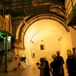 Several visitors in hard hats stand in a large underground dimly lit room at the entrance to a large circular tunnel