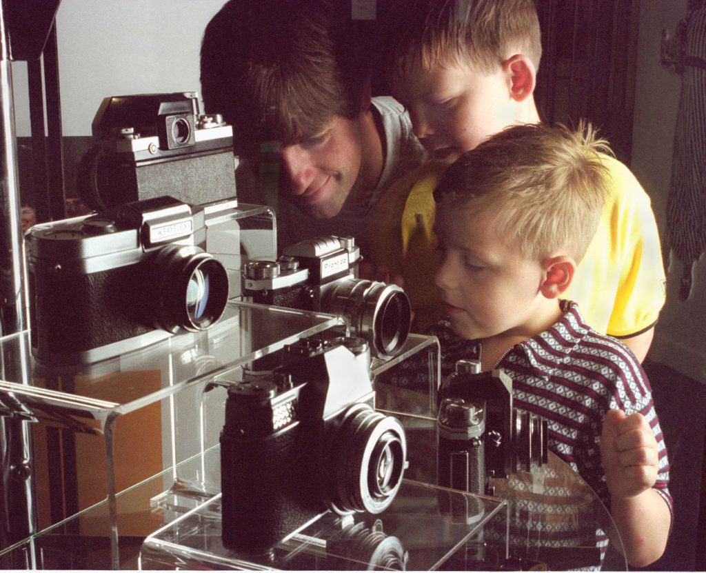 Colour photograph of a man and his two boys looking into a glass case display of cameras