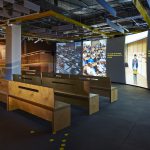 Part of the Collider exhibition showing several wooden benches in front of a large television screen showing the exhibition introductory video