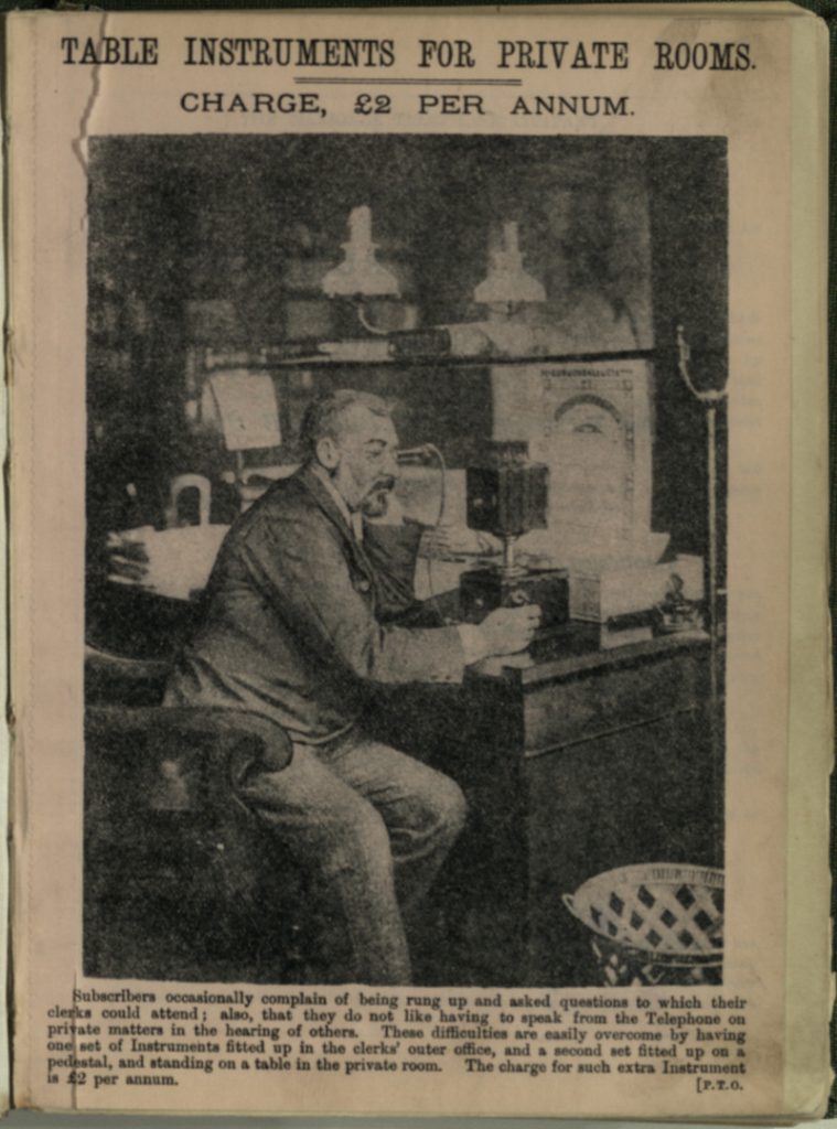 Black and white printed photograph of a man sitting at a desk holding an early table telephone from 1891