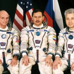 Colour photograph of three seated astronauts in space suits one of which is Dennis Tito the first space tourist