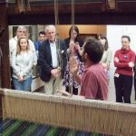 Colour photograph of of a weaver demonstrating how to work a loom in front of a museum audience