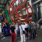 Several visitors listen to a CERN staff member talk in front of a large circular metal girder construction