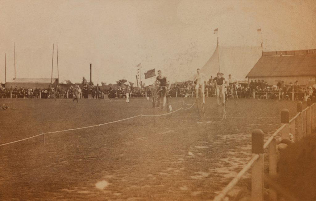 Sepia photograph of cyclists on high wheeler bicycles during a race with spectators in the background from late nineteenth century
