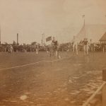 Sepia photograph of cyclists on high wheeler bicycles during a race with spectators in the background from late nineteenth century