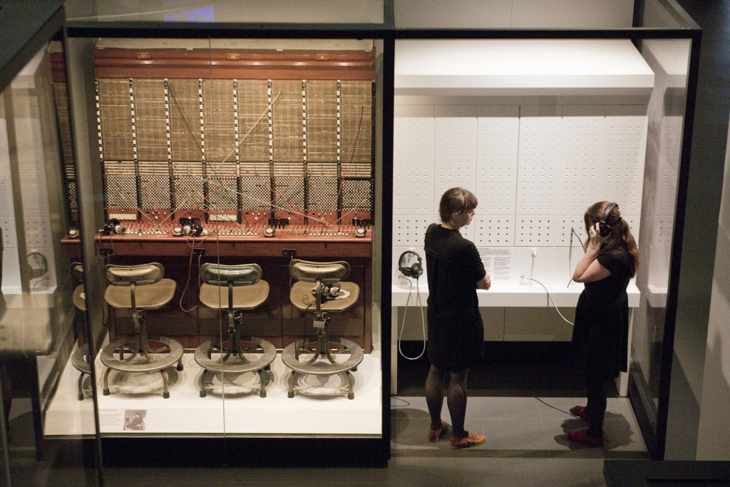Colour photograph from a high elevation of an old genuine manual telephone exchange desk and two visitors using the adjacent switchboard demonstration console