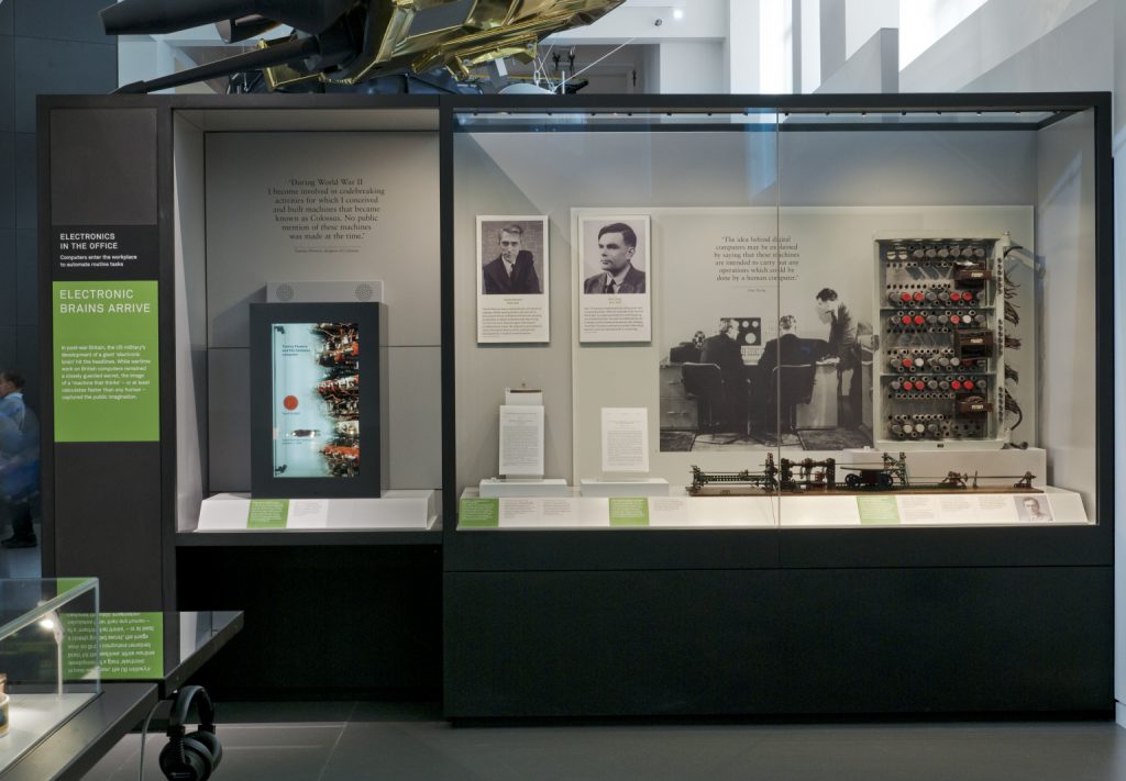 Colour photograph of a display case on computing in the Information Age exhibition showingpictures of early computer specialists a valve from an early computer and a part of the Manchester Mark 1 machine