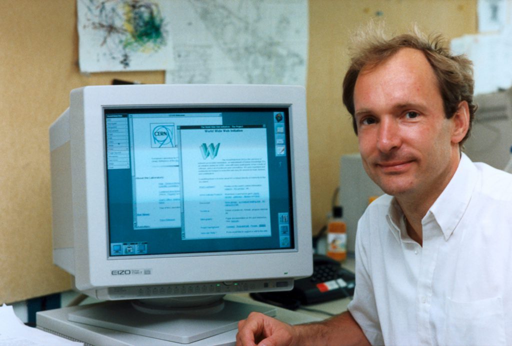 Photograph of Tim Berners-Lee sitting next to a computer display on which the words world wide web initiative are legible as well as an old CERN logo