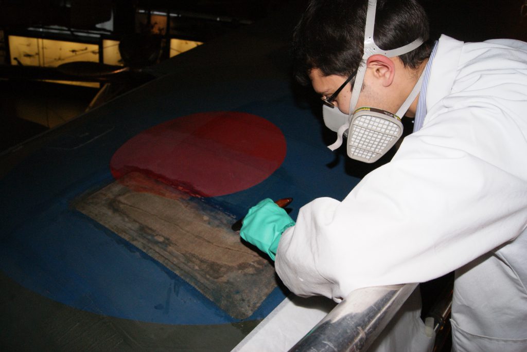 Colour photograph of a conservator doping a new patch into place to stabilise a tear in the upper-side of the Hawker Hurricane