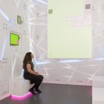 Colour photograph of the inside of a colourful and brightly lit installation in the Information Age exhibition showing a woman looking at a large screen with the text welcome to the web