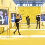 Colour photograph of visitors inside a large gallery space with photographs displayed on easels