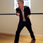 Colour photograph of Mat Fraser singing into the microphone during a performance of Cabinet of Curiosities