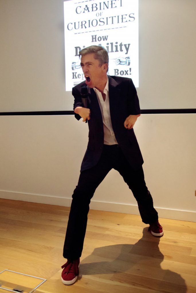 Colour photograph of Mat Fraser singing into the microphone during a performance of Cabinet of Curiosities
