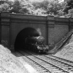 Black and white photograph of the Flying Scotsman steam train emerging from a tunnel. Anti diesel train graffiti is visible on the brickwork