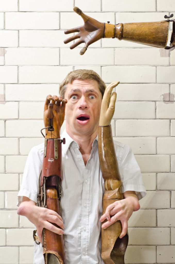 Colour photograph of Mat Fraser holding up two historical wooden prosthetic arms