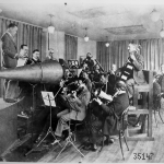 Black and white photograph of a studio orchestra playing into a megaphone shaped recording instrument