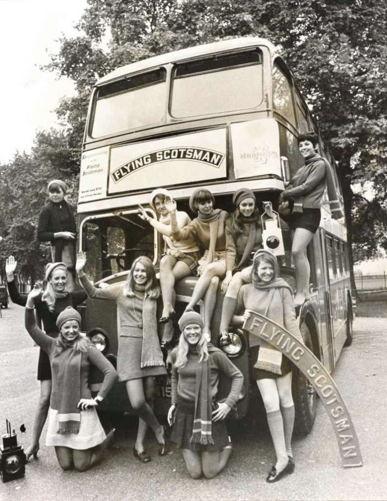 Black and white publicity photograph of a double decker bus with Flying Scotsman printed on the front and a number of young women surrounding the bus