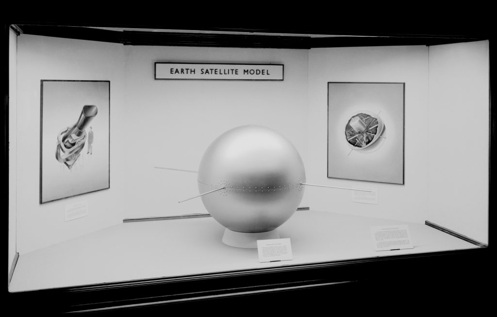 Black and white photograph of a model of an early satellite on display as part of an exhibition