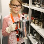 Colour photograph of a young girl holding an early metal kettle