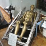 Colour photograph of a gold coloured mannequin lying inside a space craft pilot seat