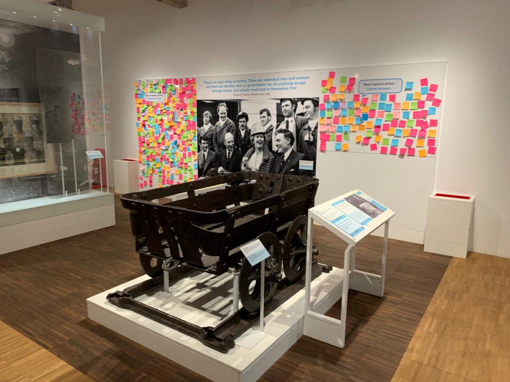Colour photograph of a coalmining cart displayed in front of Margaret Thatcher's photograph in an exhibition gallery