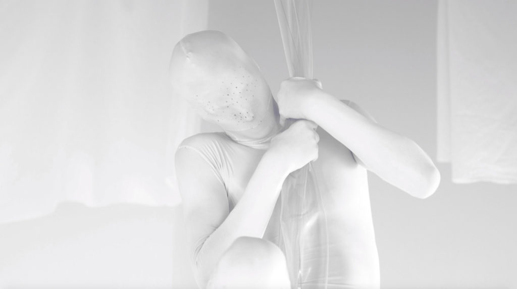 Video still showing a figure in white lycra against white fabrics