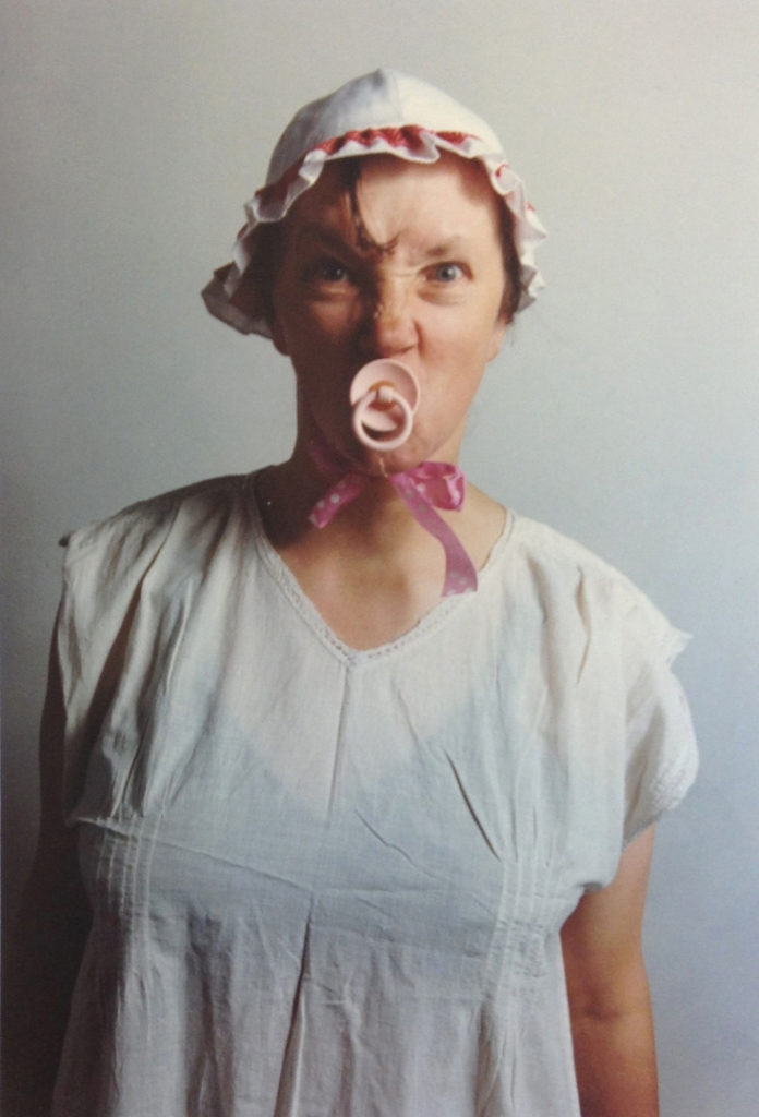 Colour photograph of Jo Spence dressed in baby clothes with dummy