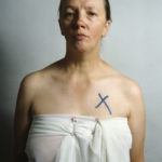 Colour photograph of Jo Spence wrapped in a sheet with a black cross over her breast
